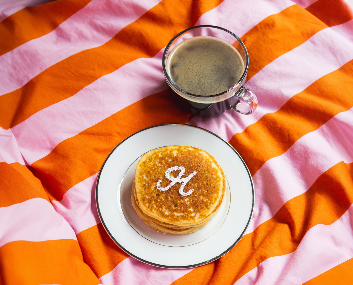 A stack of golden pancakes on a white plate with a black rim, topped with powdered sugar written in the shape of the letter 'H', set on an orange and pink striped blanket next to a glass mug of coffee.